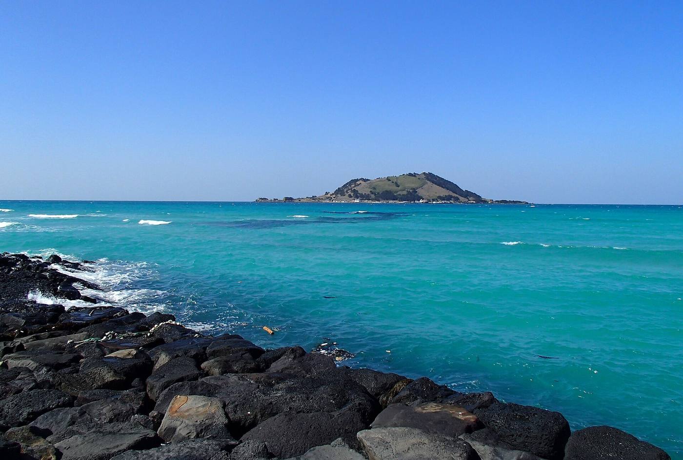 A sea of bright turquoise water seen over black volcanic rocks, with the small green island of Biyangdo on the horizon under a clear blue sky, from the shore at Hyeopjae.