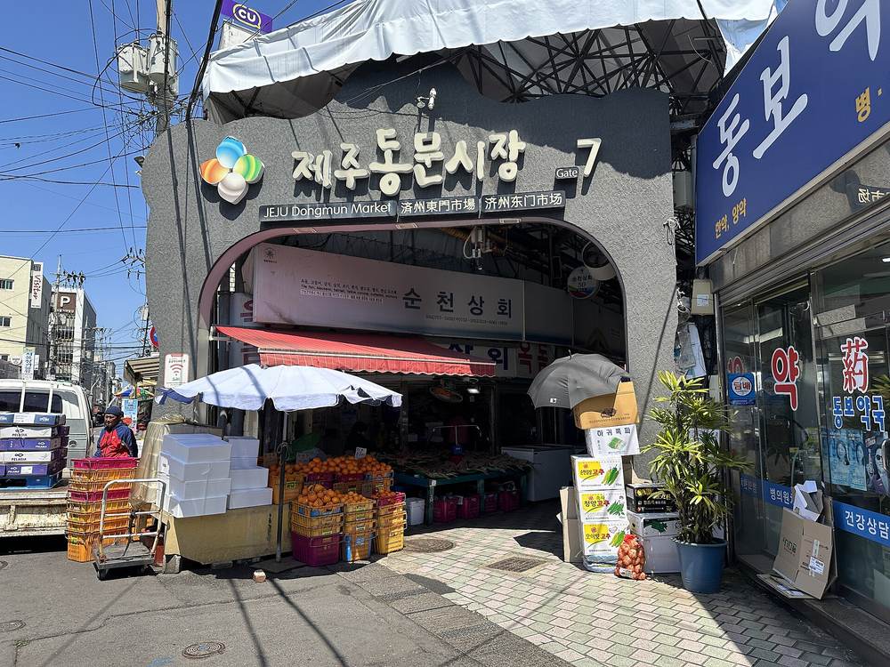 The entrance to Jeju Dongmun Market, Gate 7, a grey arched gateway with a large Korean sign reading 제주동문시장 and smaller signs in English and Chinese, with crates of tangerines and a produce stall under a red awning on a sunny street.