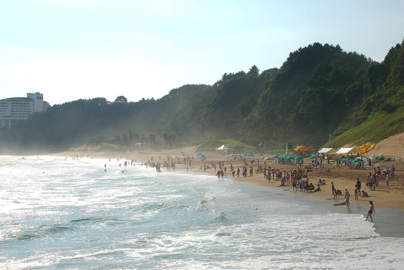 A wide sandy beach with people paddling in the surf below a steep wooded slope, and a large white hotel on the cliff top at the left, at Jungmun on Jeju's south coast.