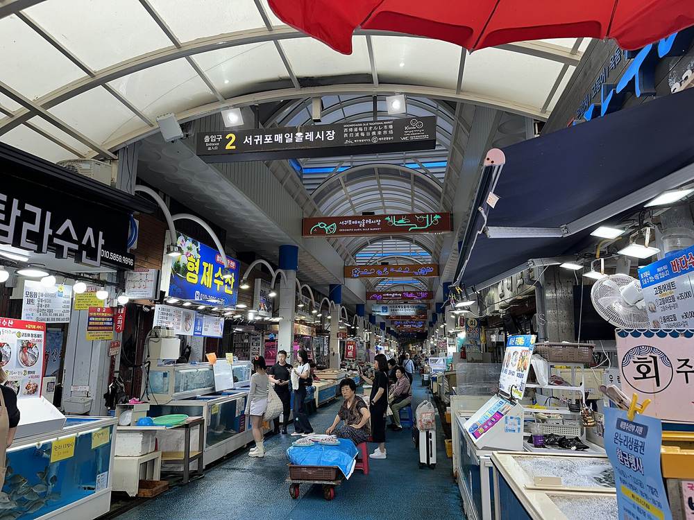 A covered market street with a curved white roof, a hanging sign reading 서귀포매일올레시장 with the gate number 2, fish tanks and stalls on both sides, and shoppers walking on a blue-painted floor.