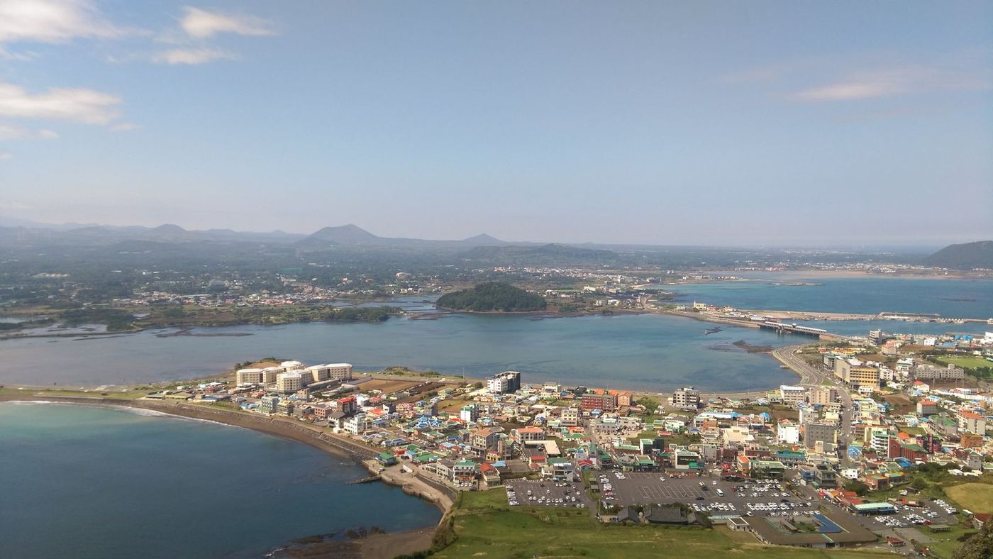 The village of Seongsan seen from high above: a cluster of low colourful buildings and a car park between two bays, a small wooded hill in the lagoon, a causeway, and the hills of eastern Jeju behind.