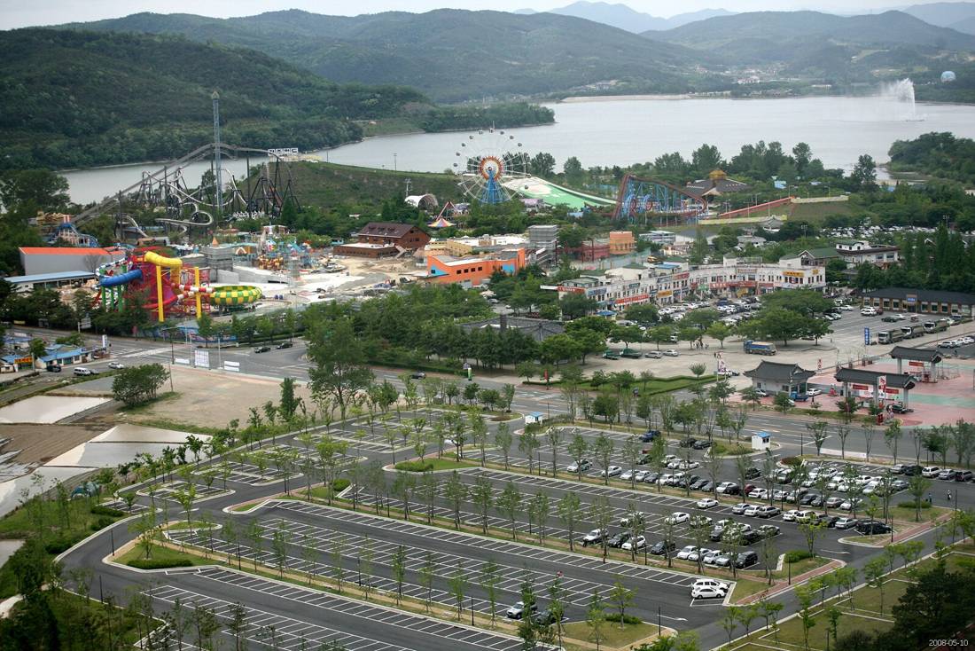 An aerial view of Bomun Lake Resort in Gyeongju in May 2008, with a large car park in the foreground, the Gyeongju World amusement park with a roller coaster and Ferris wheel behind it, and the lake with a fountain and wooded hills in the distance.