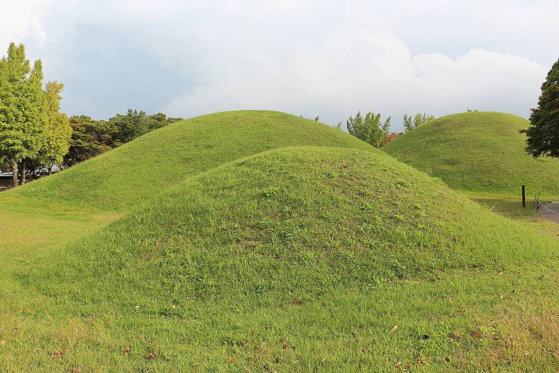 Grassy royal tomb mounds of the Daereungwon complex in Gyeongju under a cloudy sky, with trees on the left and a small fence post on the right.