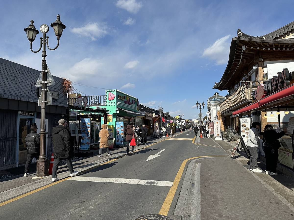 The main street of Hwangnidan-gil in Gyeongju in January 2025, with a two-lane road, ornate street lamps, a timber hanok-style shop on the right, a green-fronted snack shop on the left and visitors in winter coats on the pavements.