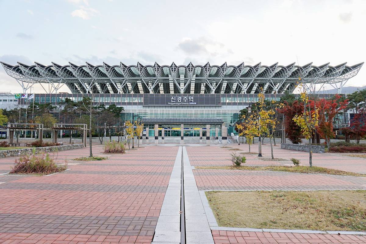 The front of the KTX station in Gyeongju in November 2019, when it was still named Singyeongju, showing the white steel canopy shaped like rows of roof tiles above a glass hall with the sign 신경주역, and a paved plaza with autumn trees.