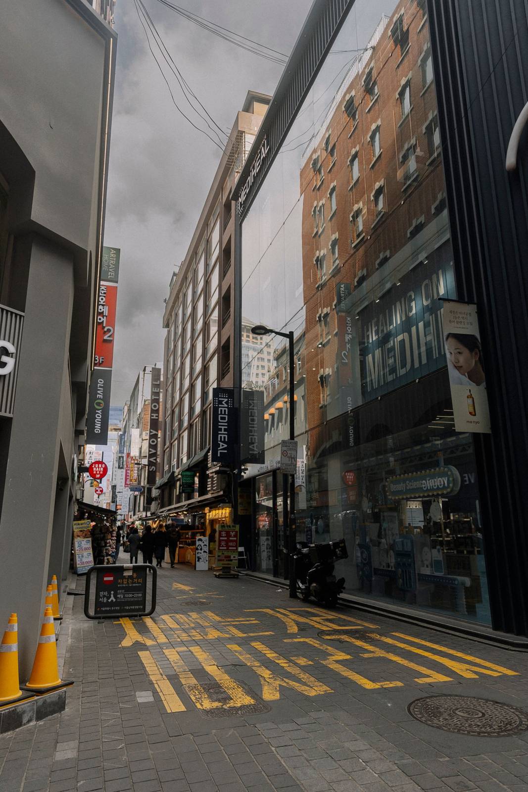 A Myeongdong side street on an overcast day: a Mediheal store on the right, an Olive Young sign on the left, the Savoy Hotel sign and street stalls further down, and a currency-exchange board showing dollar, yen, yuan and Hong Kong dollar signs.