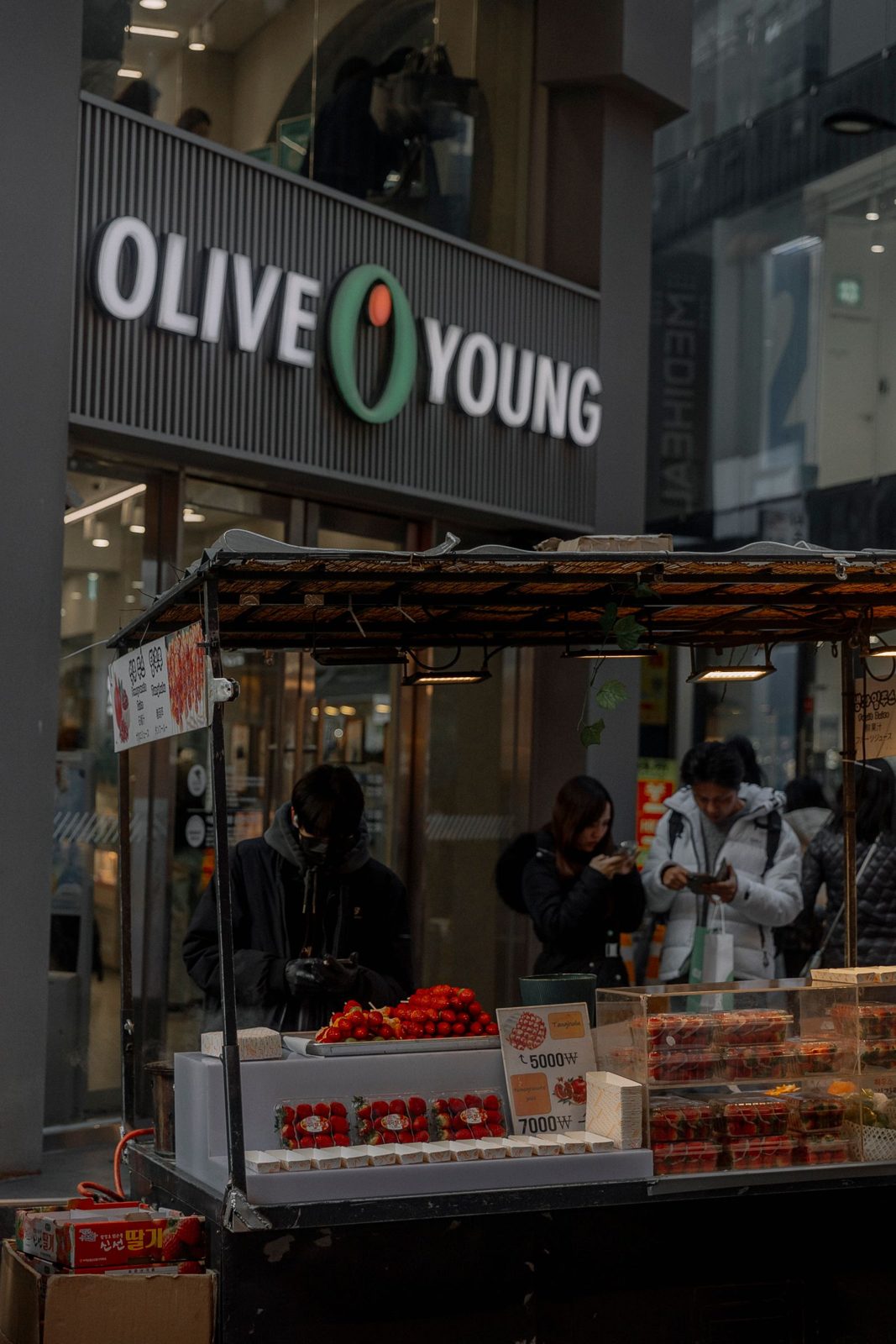 An Olive Young store sign above a street cart selling strawberry tanghulu, with a price board showing ₩5,000 and ₩7,000 and shoppers looking at their phones.