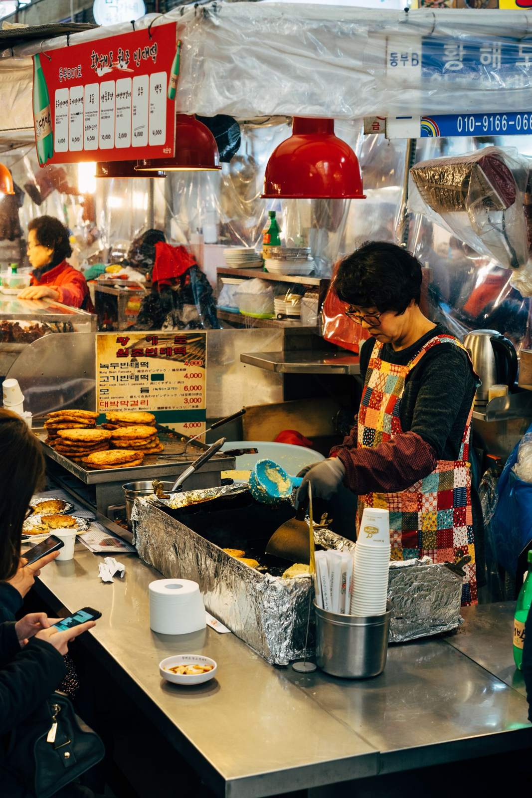 A market stall frying mung-bean pancakes, with a menu board reading 녹두빈대떡 ₩4,000, 고기빈대떡 ₩4,000, 모듬 ₩6,000 and 대박막걸리 ₩3,000, and customers at the counter on the left.