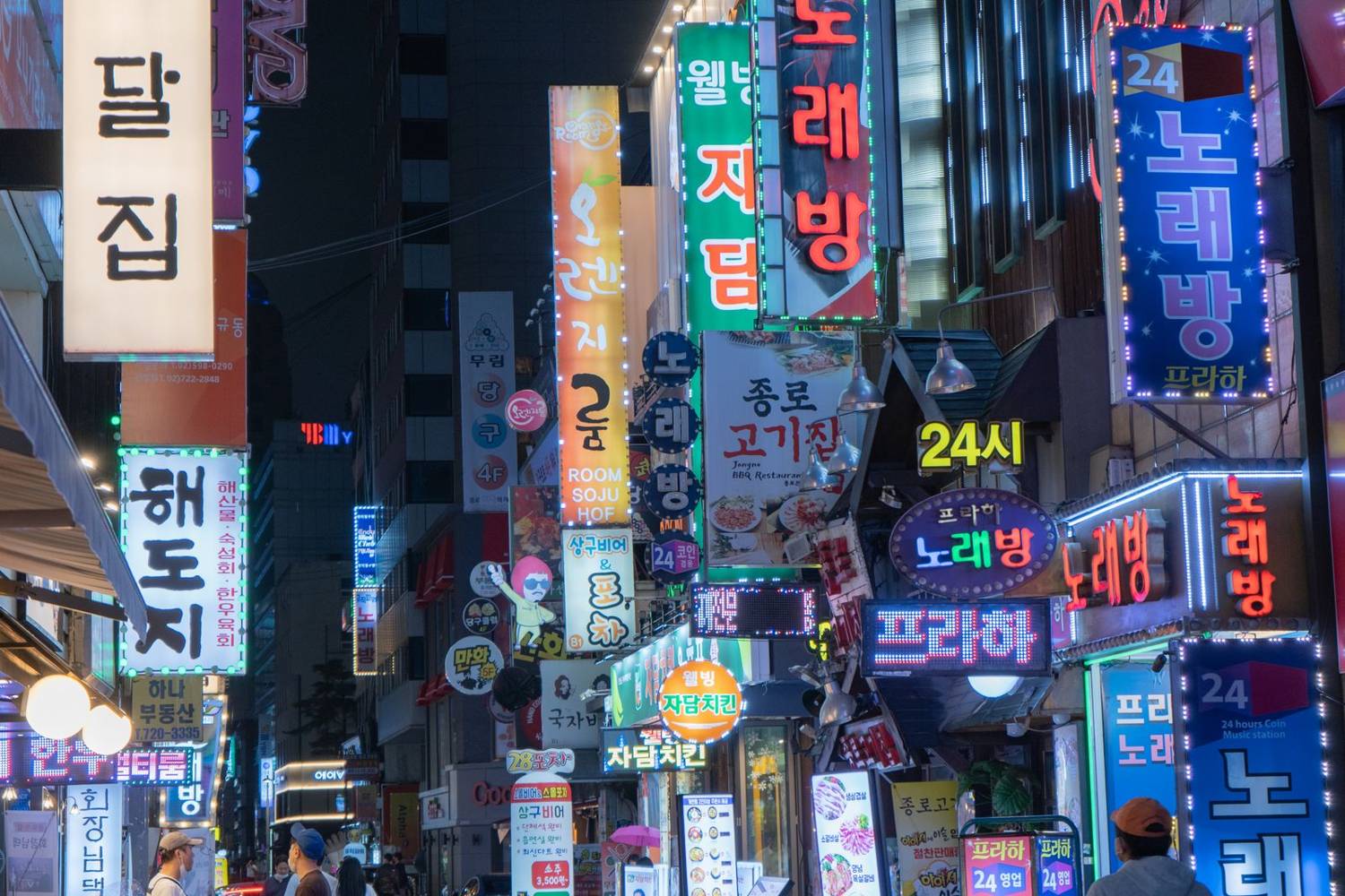 Vertical neon signs for restaurants, pubs and noraebang karaoke rooms on a Seoul side street at night, with a standing board in front of a pub advertising soju from ₩3,500.