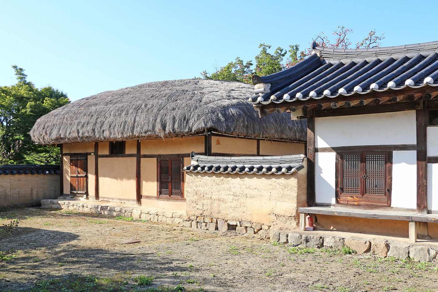 A traditional house in Hahoe Village: a thatched building with earth-coloured walls beside a tile-roofed wing with white plaster walls and wooden lattice doors, behind a bare earth yard.