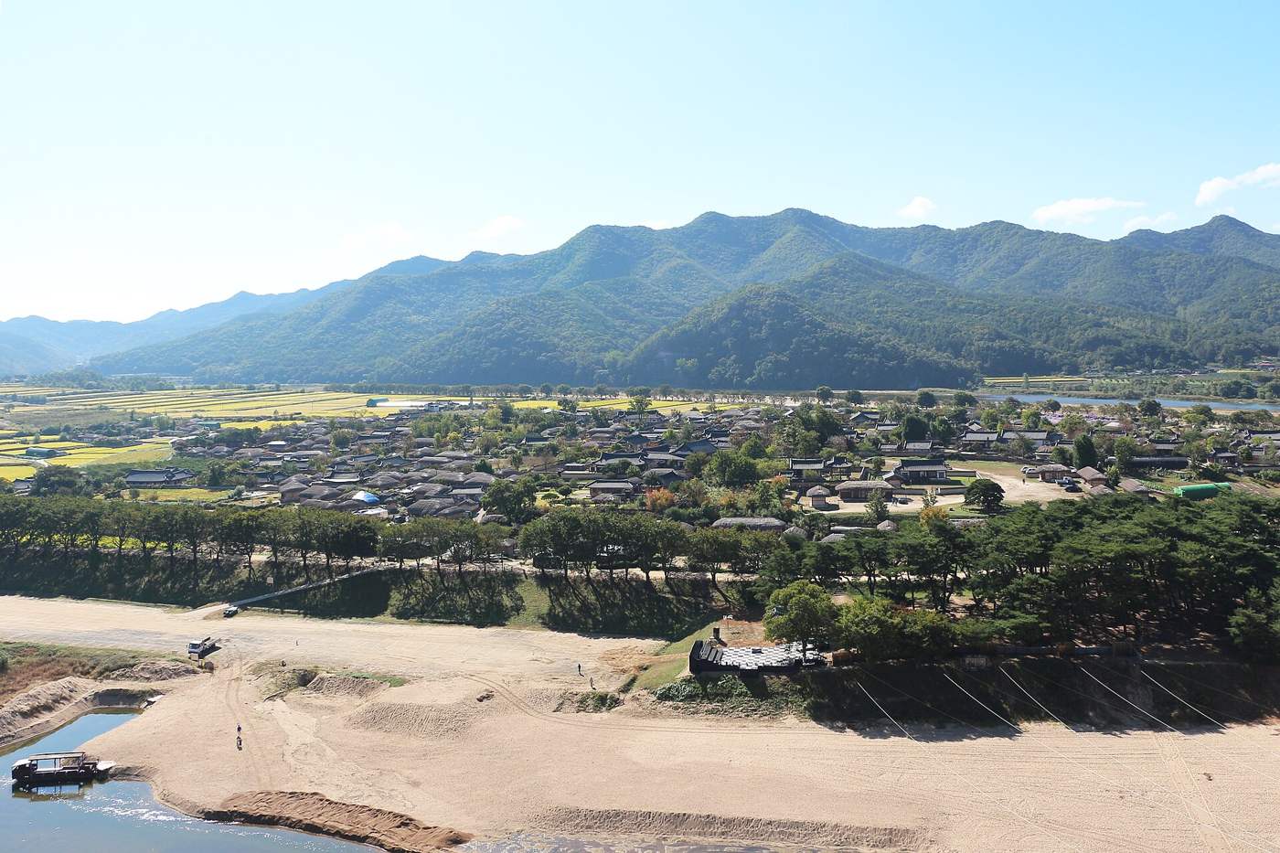 The thatched and tile-roofed houses of Hahoe Village under a blue October sky, seen across a sandy bend of the Nakdong River, with a pine grove along the bank and mountains behind.