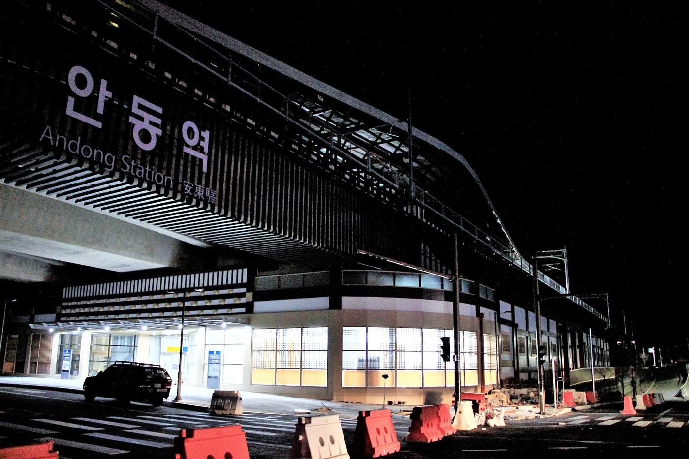 Andong Station at night: a long canopy of slatted wood over the entrance, with the station name 안동역 and Andong Station in white letters above glass doors.