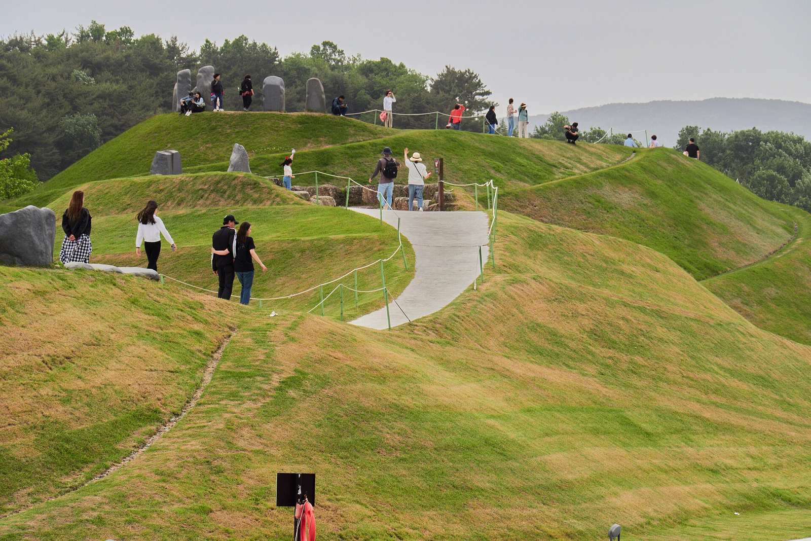 Visitors walking up a paved path that winds around grassy, sculpted mounds topped with standing stones in Suncheon Bay National Garden.