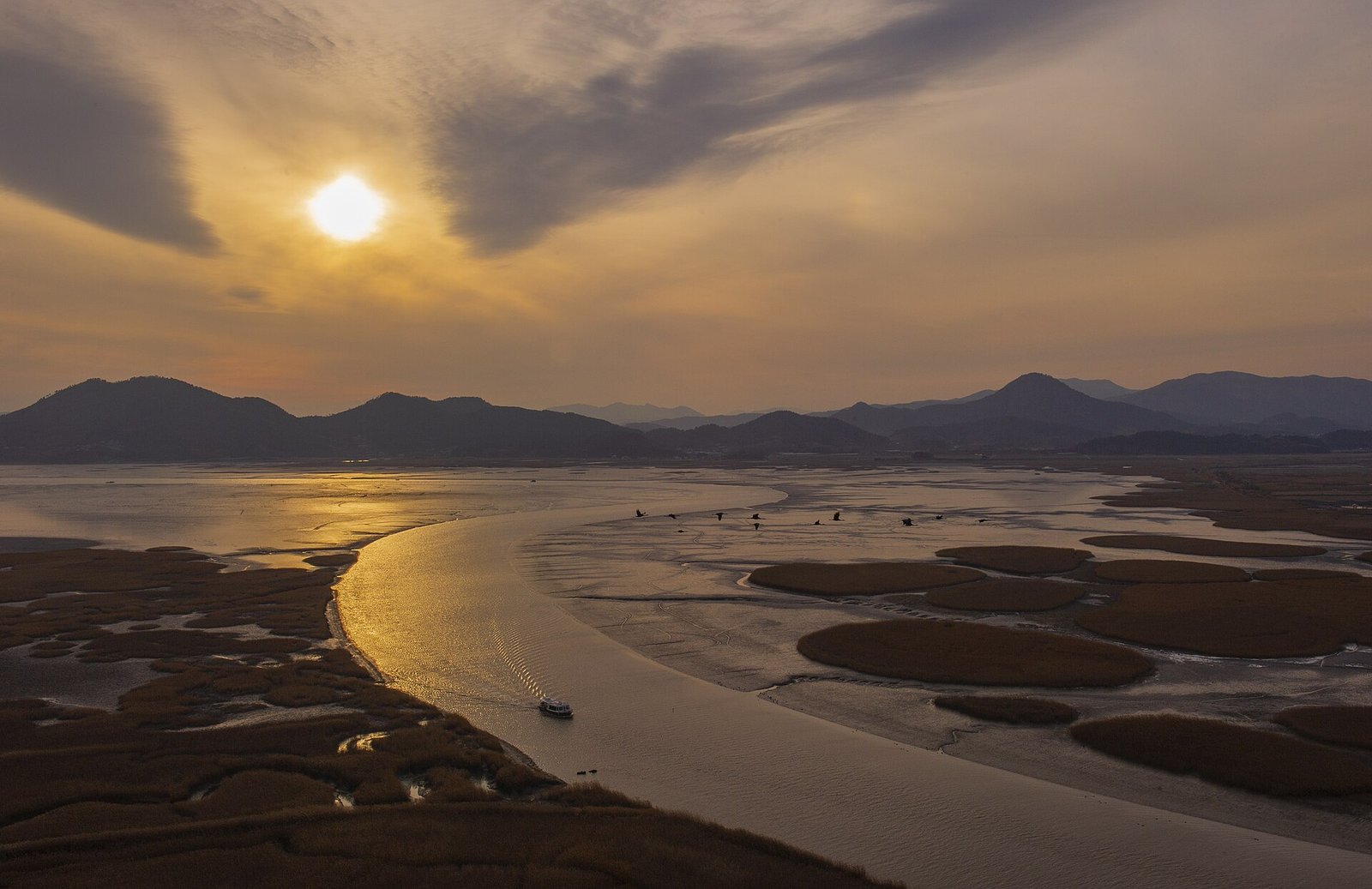 Sunset over Suncheon Bay: the sun low behind clouds, a curving tidal channel lit gold, round reed islands on dark mudflats and a small boat on the water, January 2016.