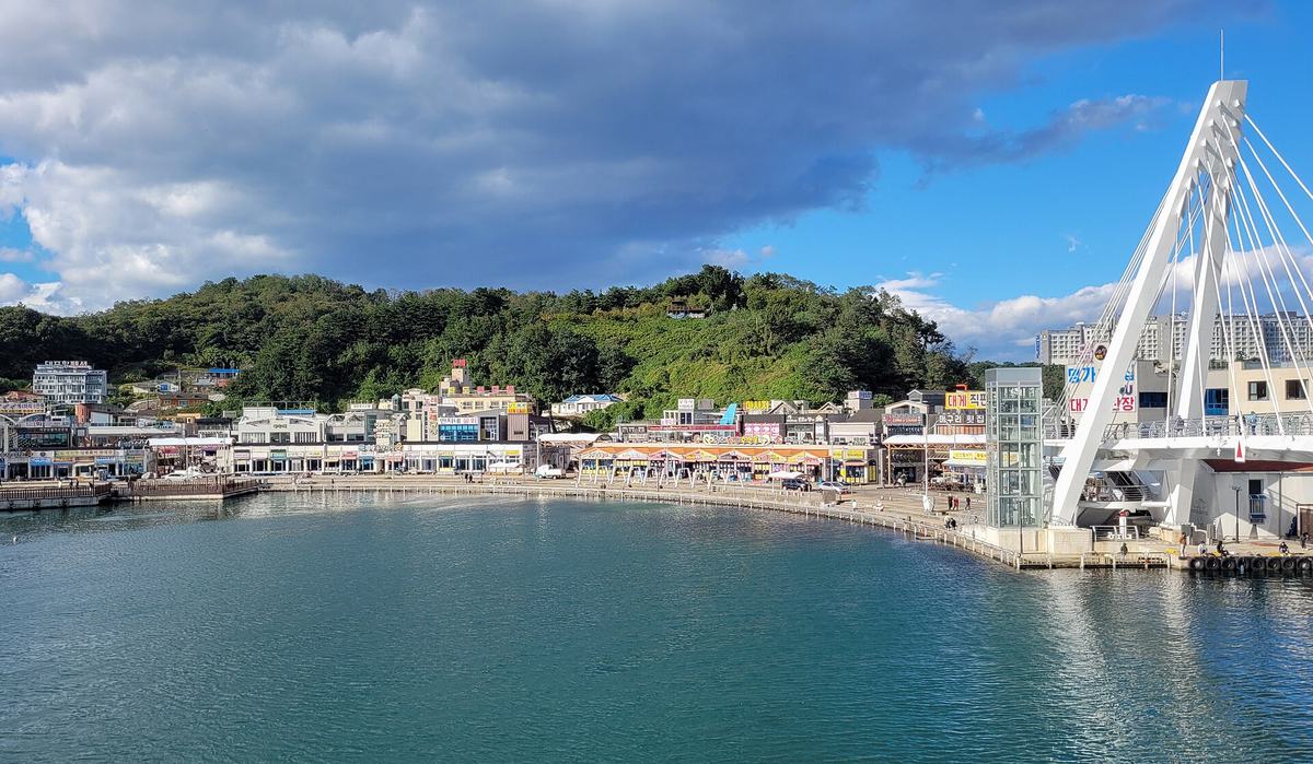 Daepo Port in Sokcho on a sunny day in October 2022, with calm blue-green water in the foreground, a curved quay lined with low seafood restaurants, a white cable-stayed pedestrian bridge on the right and wooded hills behind.