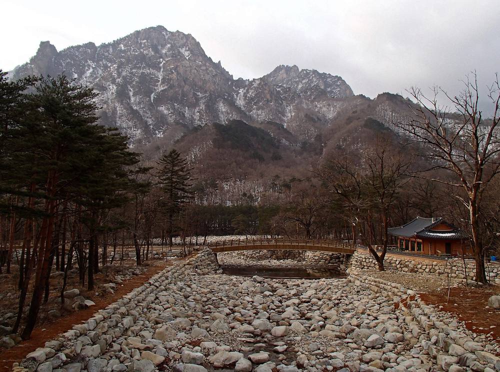 The area near the entrance of Sinheungsa temple in Seoraksan National Park in early March 2013, with a dry stone-lined stream bed and a small stone bridge in front of a traditional temple building, bare trees, and rugged rocky peaks with patches of snow behind.