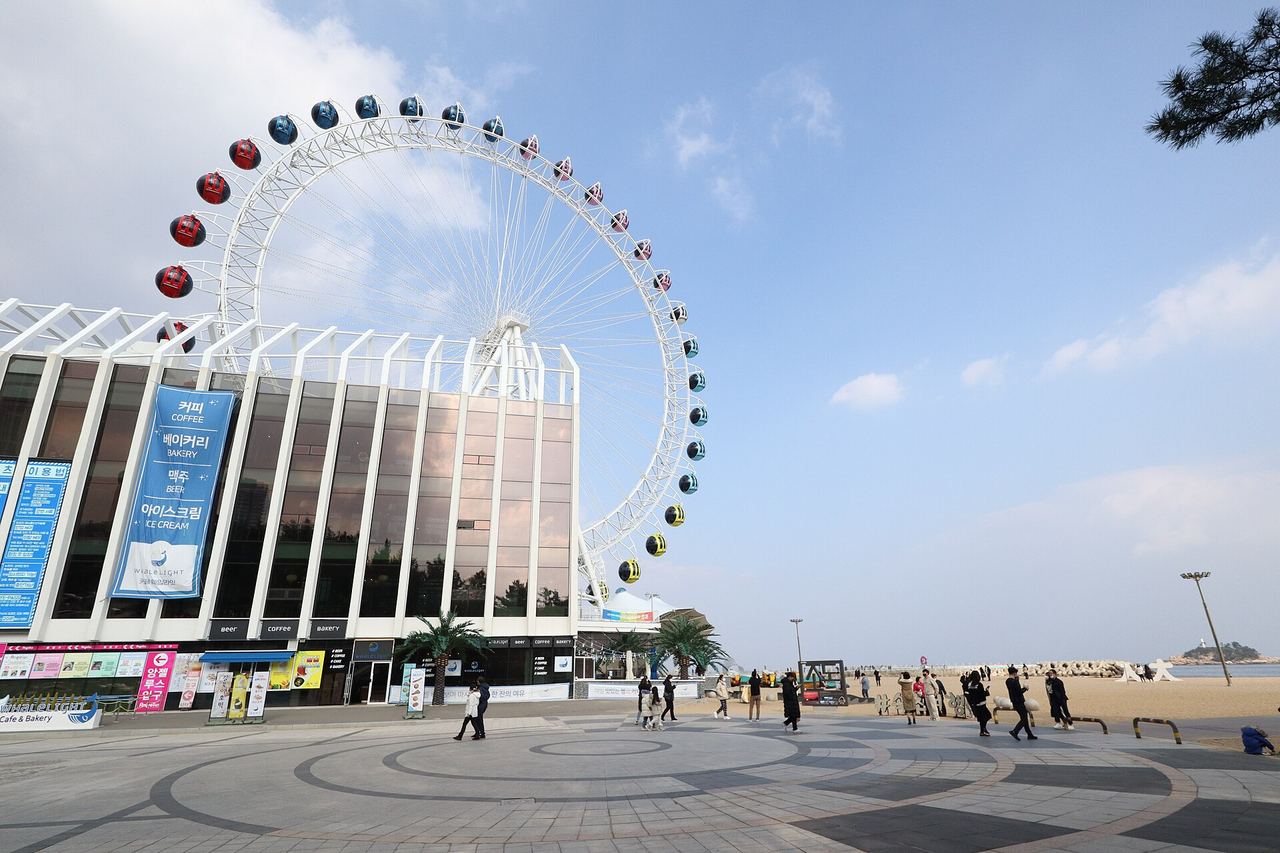 The Sokcho Eye, a large white Ferris wheel with red and blue gondolas, beside a glass-fronted building on the paved promenade at Sokcho Beach in December 2022, with people walking and the sea and a small island on the right.