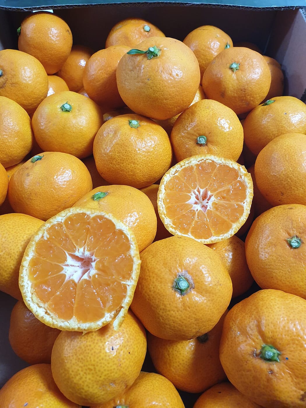 A box of fully orange, thin-skinned mandarins with green stem ends, two of them cut in half to show the segments.