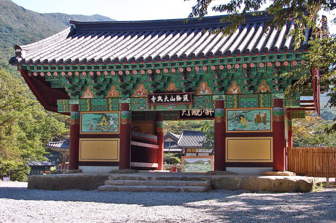 The front gate of Daeheungsa temple in Haenam, Korea: a single-storey gate with a curved tiled roof, green, red and gold painted brackets, red pillars, murals on the side panels and a plaque reading 頭輪山大興寺, with forested mountains behind, in October 2011.