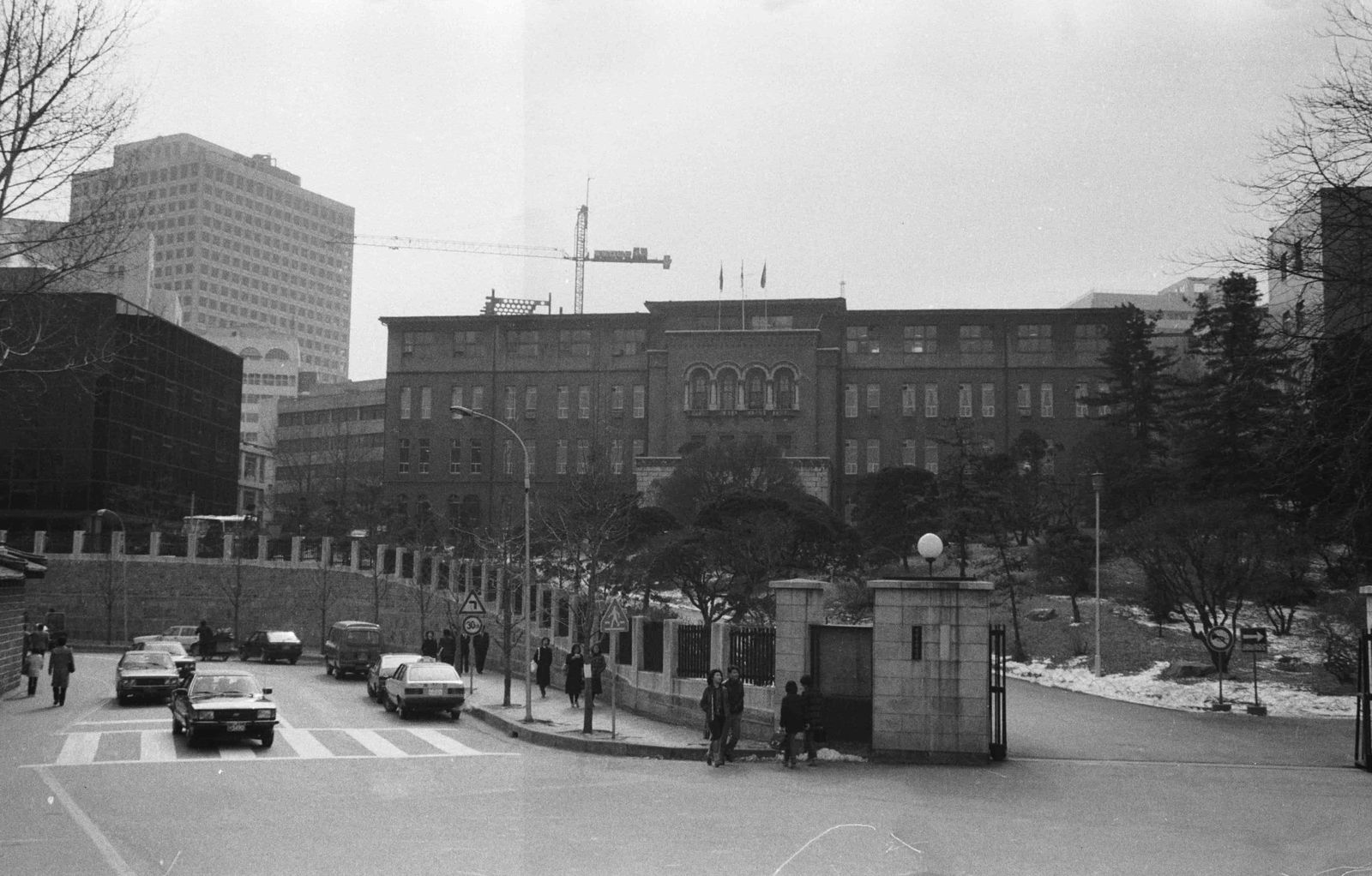 A black-and-white photograph of the old Supreme Court of Korea building at Seosomun-dong, Seoul, on 10 February 1983: a long brick building with an arched central entrance behind a stone gate, cars at a crossing in front, snow on the bank to the right and office towers behind.