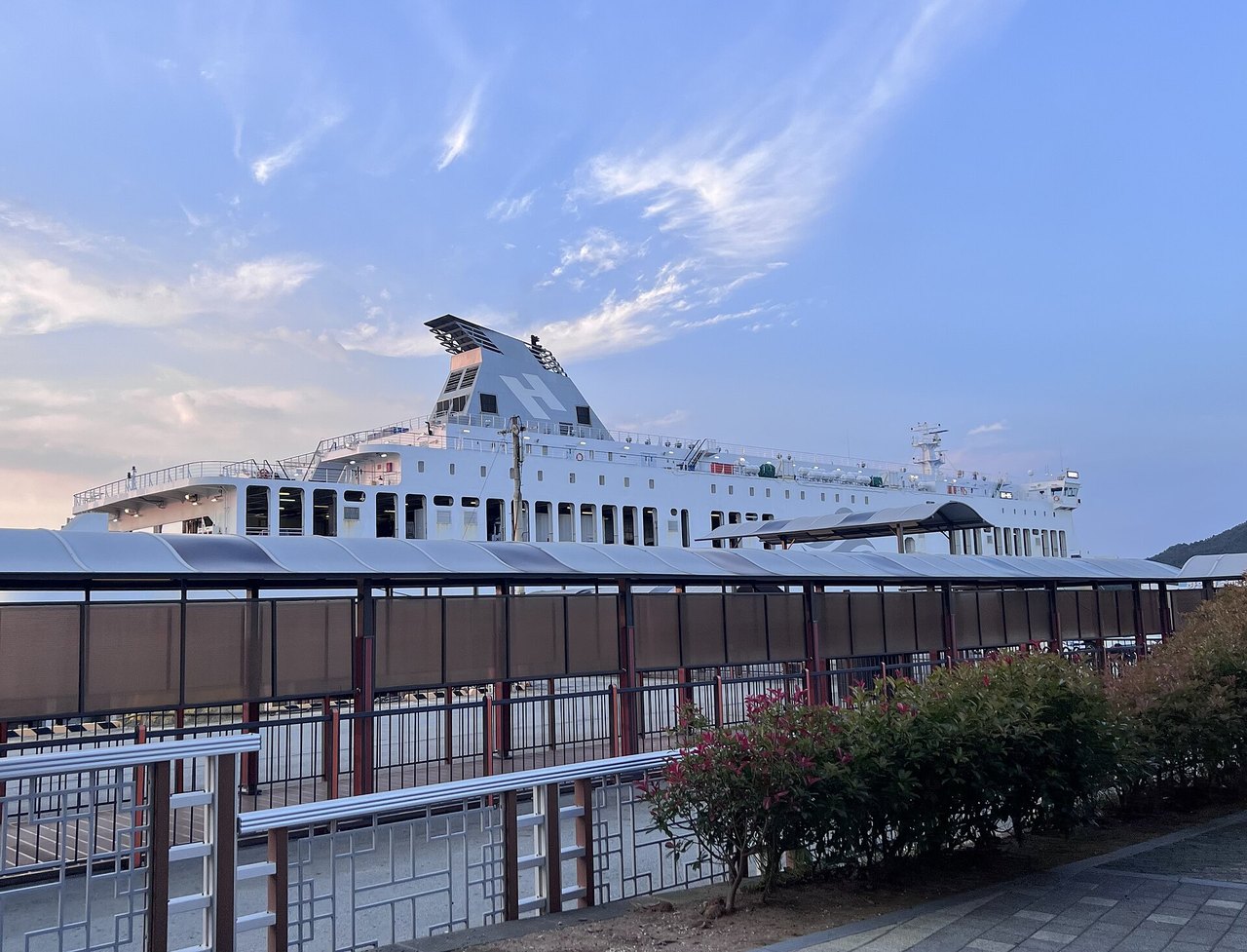 A large white car ferry with a grey funnel marked with a big white letter H, moored behind a covered walkway and railings at a ferry terminal in Wando, under a blue evening sky with wispy clouds, 10 June 2026.
