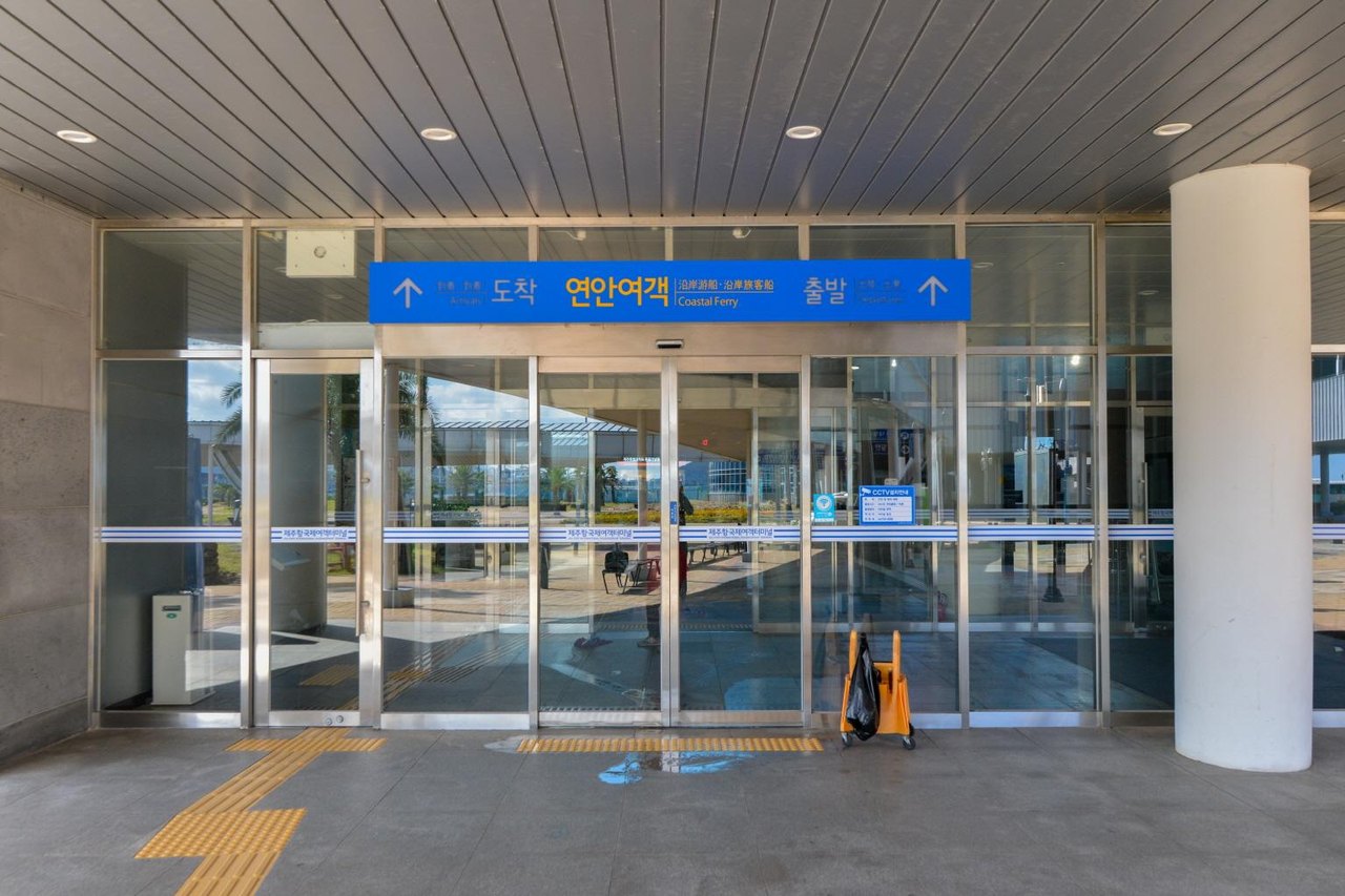 The covered entrance to the coastal ferry hall at Jeju Port, with a blue overhead sign reading 도착 연안여객 출발 and "Coastal Ferry" in English, glass sliding doors with a strip reading 제주항국제여객터미널, and tactile paving on the floor, October 2019.