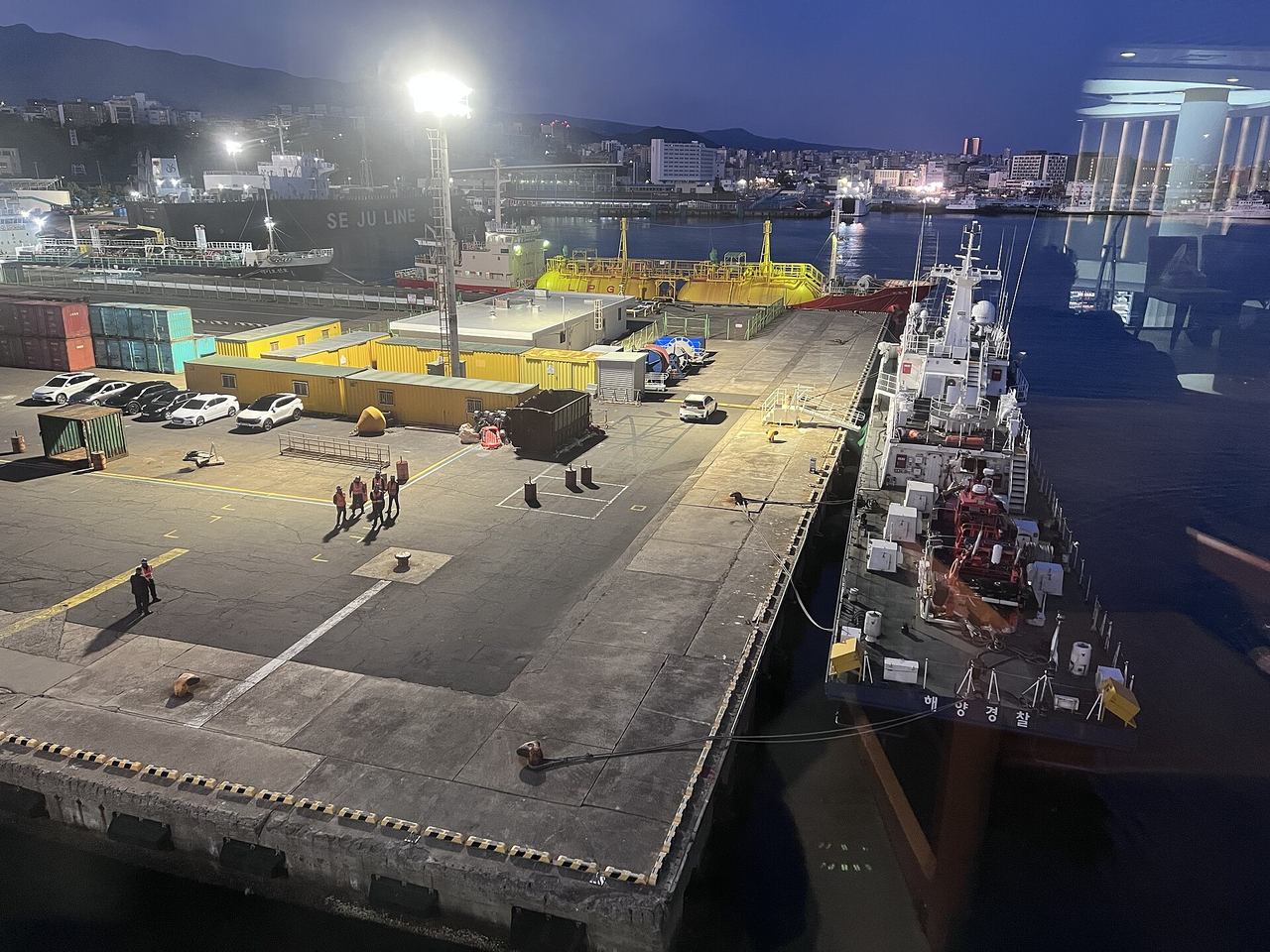 Jeju Port at dawn seen through a ferry window from high above the quay, with floodlit concrete piers, parked cars and yellow site cabins, a Korea Coast Guard ship marked 해양경찰 moored alongside on the right, and Jeju City's lights and hills behind, 8 April 2026.