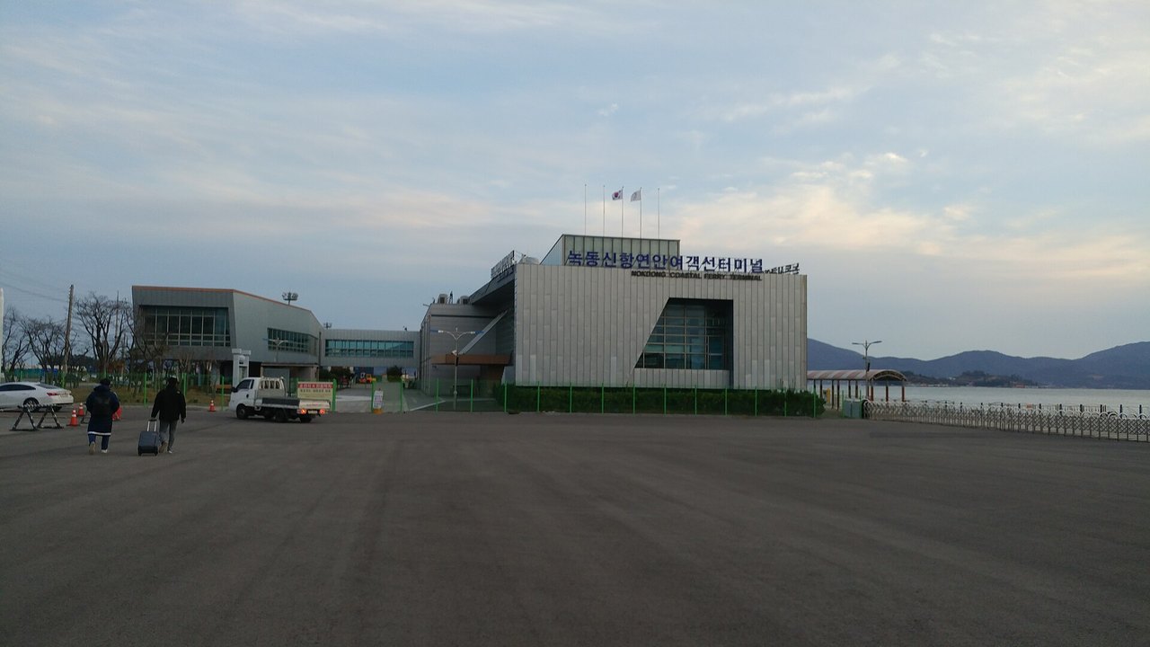 A low, modern grey ferry terminal with a sign reading 녹동신항연안여객선터미널 and "NOKDONG COASTAL FERRY TERMINAL" on the roof, a wide empty car park in front with two people pulling a suitcase, and the sea and hills to the right, December 2019.