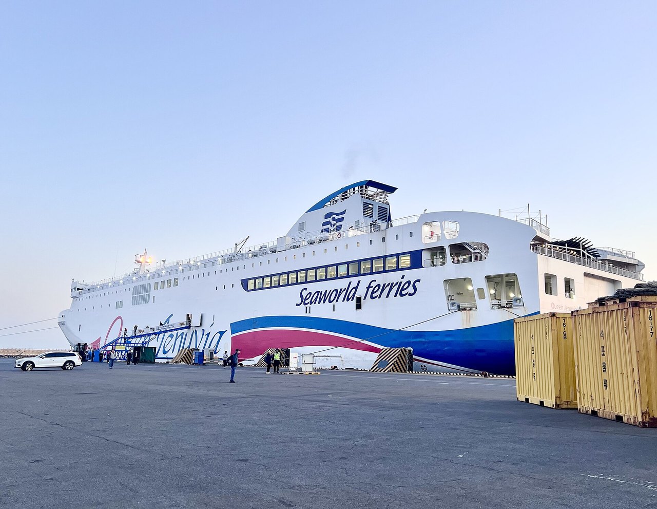The white Seaworld Ferries car ferry Queen Jenuvia, with its name in pink and blue script on the hull and "Seaworld ferries" on the side, moored at a quay in Jeju Port at dawn, with a few passengers and staff on the tarmac and yellow cargo containers to the right, 8 April 2026.
