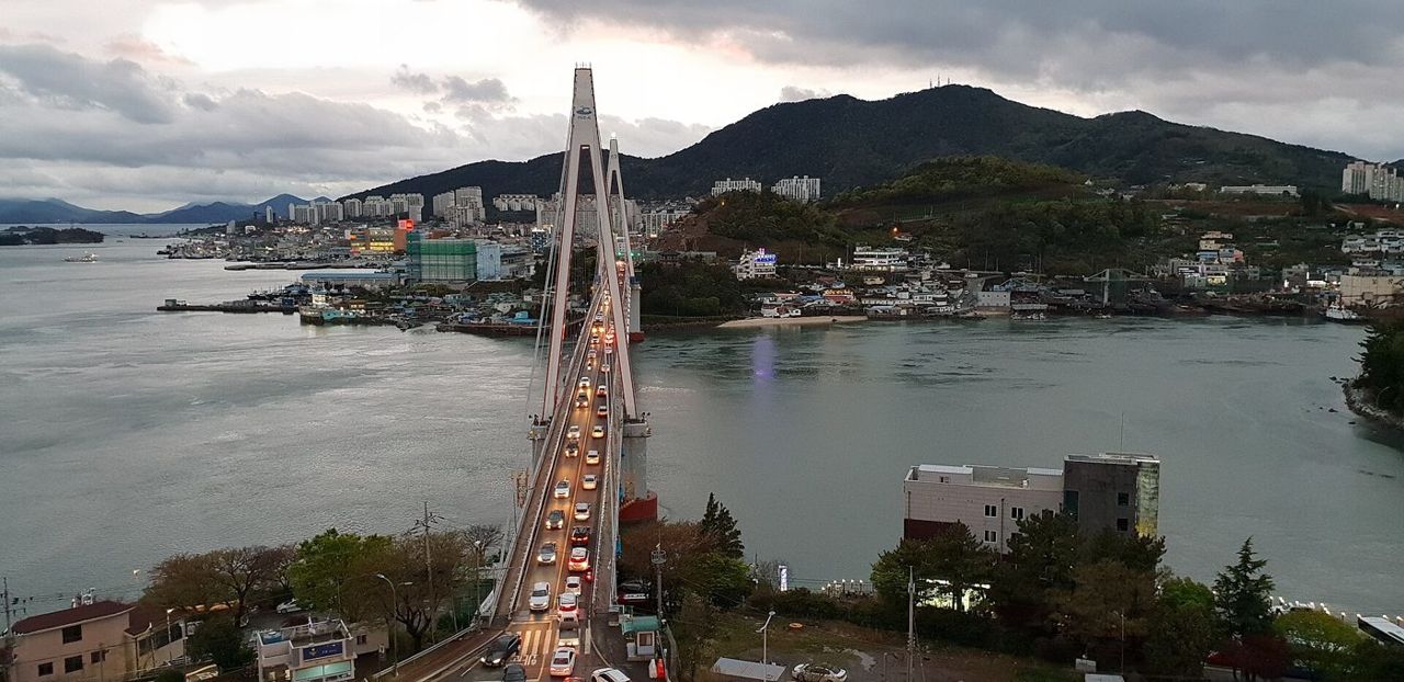 The white cable-stayed Dolsan Bridge seen from Dolsan Park in the evening, with car headlights on the deck, the water of the strait and the hillside city of Yeosu behind under a grey sky, April 2018.