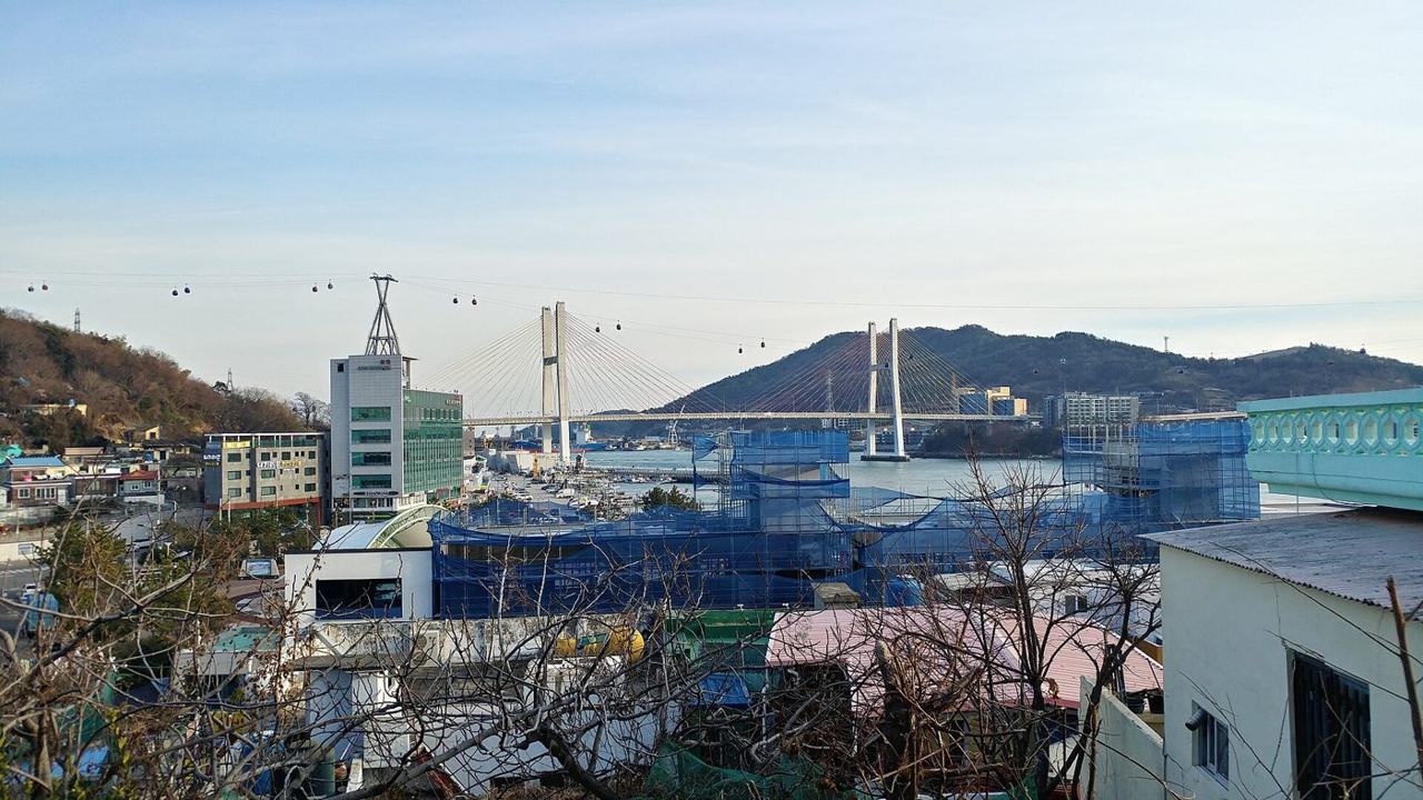 The white cable-stayed Geobukseon Bridge in Yeosu seen from a hillside in Jonghwa-dong, with a line of small cable-car cabins crossing the sky above it, rooftops and buildings wrapped in blue construction netting in the foreground, January 2020.