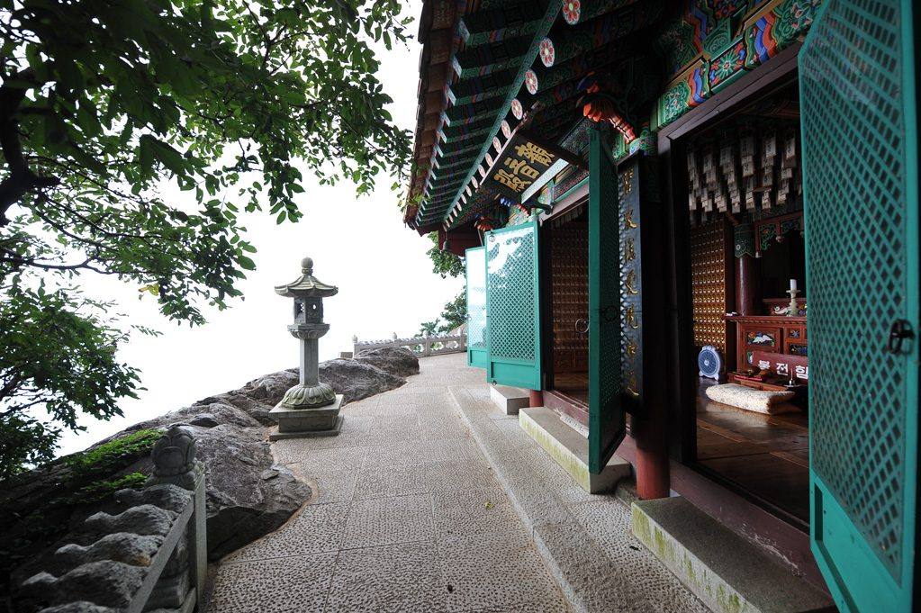 A paved path along the cliff edge at Hyangiram hermitage, with a stone lantern and a large boulder on the left, an open hall with green latticed doors and painted eaves on the right, and a hazy white sky beyond the cliff edge, August 2010.