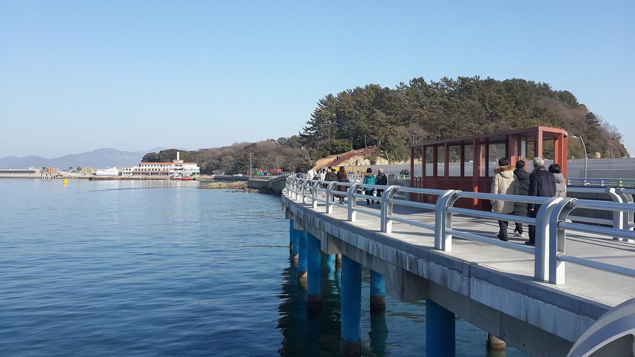 A white railed walkway over calm blue water leading towards the wooded island of Odongdo, with people walking and a red shelter on the path, under a clear sky in January 2019.