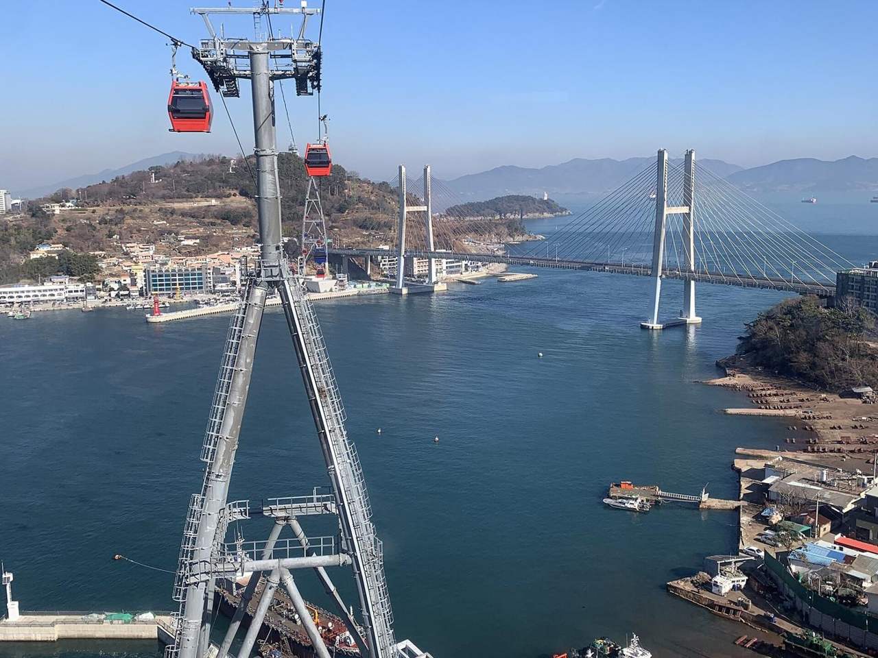 Two red cabins of the Yeosu Maritime Cable Car on the cable beside a grey steel pylon, high above blue harbour water, with the white twin-towered Geobukseon Bridge and the hills of Dolsan behind, January 2022.