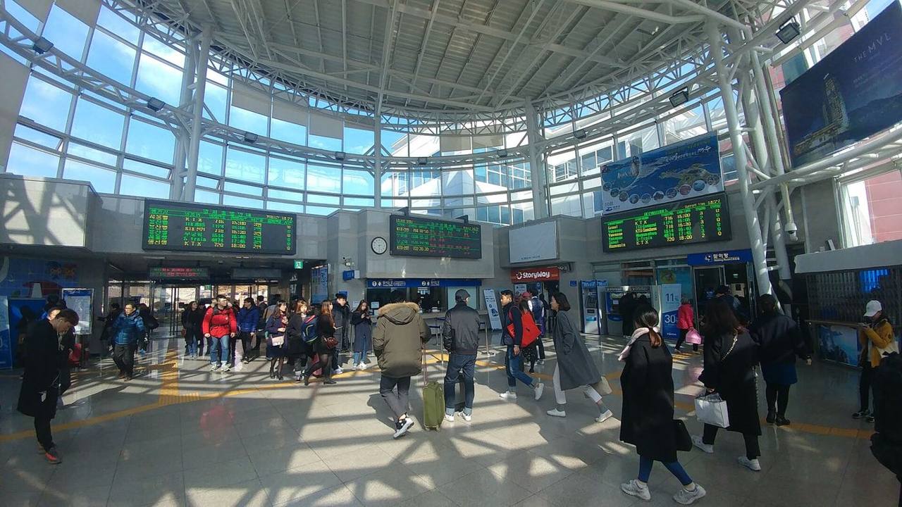 The bright concourse of Yeosu EXPO Station under a curved white glass roof, with departure boards above the ticket gates and travellers with coats and luggage walking across the floor, February 2017.