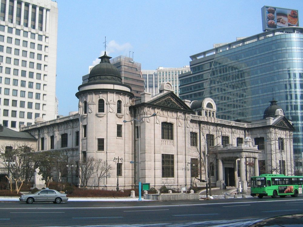 The Bank of Korea Money Museum on Namdaemun-ro, Seoul: a grey granite building with a domed corner tower and arched windows, a green city bus in front and office towers behind.