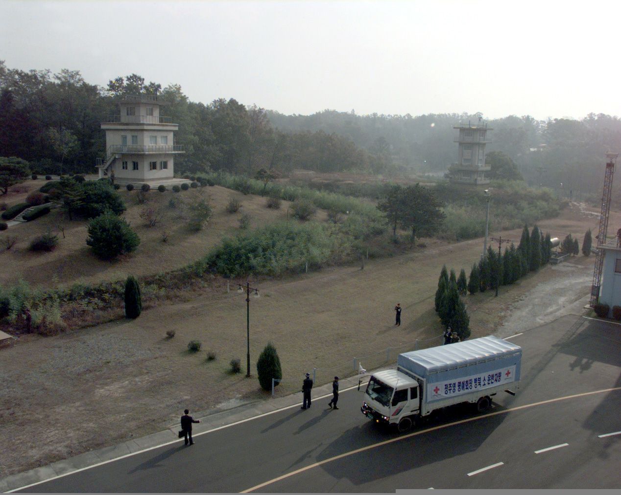 A white truck with Red Cross marks and a Korean banner drives along the road at Panmunjom, past two concrete watchtowers on a dry autumn hillside, with security staff beside the road.