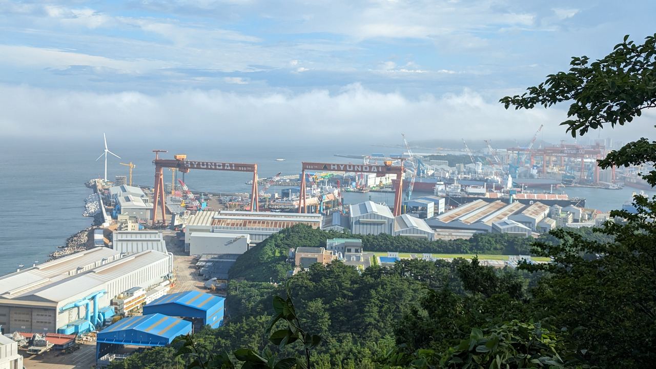 HD Hyundai Heavy Industries' Ulsan shipyard seen from a wooded hill on the East Sea coast: red gantry cranes marked HYUNDAI over the docks, factory sheds along the shore and a wind turbine on the horizon.