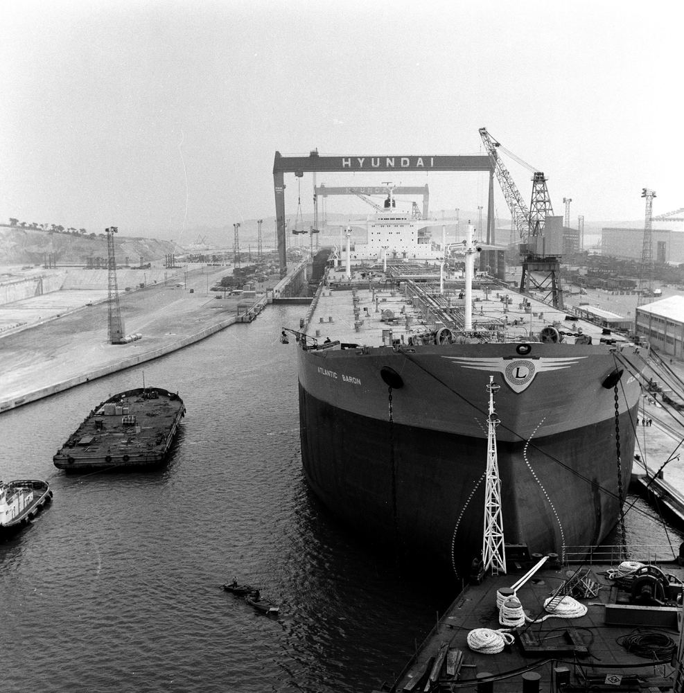 A black-and-white photograph of Hyundai's Ulsan shipyard: a huge tanker with ATLANTIC BARON and an L crest painted on its bow sits in a fitting-out basin, a gantry crane marked HYUNDAI stands behind it, and small barges float in the foreground.
