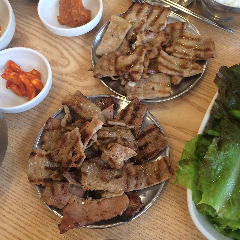 Two round stainless-steel plates piled with grilled slices of pork showing dark grill marks, with small bowls of red pickled radish and dipping paste and a heap of lettuce leaves at the edge, July 2016.