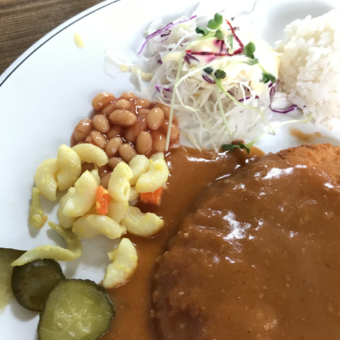 A Korean-style pork cutlet covered in glossy brown sauce on a white plate, with macaroni salad, baked beans, pickle slices, shredded cabbage with dressing and a scoop of rice, May 2017.