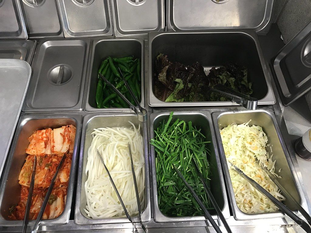 A refrigerated self-service counter of stainless trays holding kimchi, white radish strips, garlic chives, cabbage salad with corn, green chillies and lettuce, with tongs in each tray, at a Seoul drivers' restaurant, April 2017.