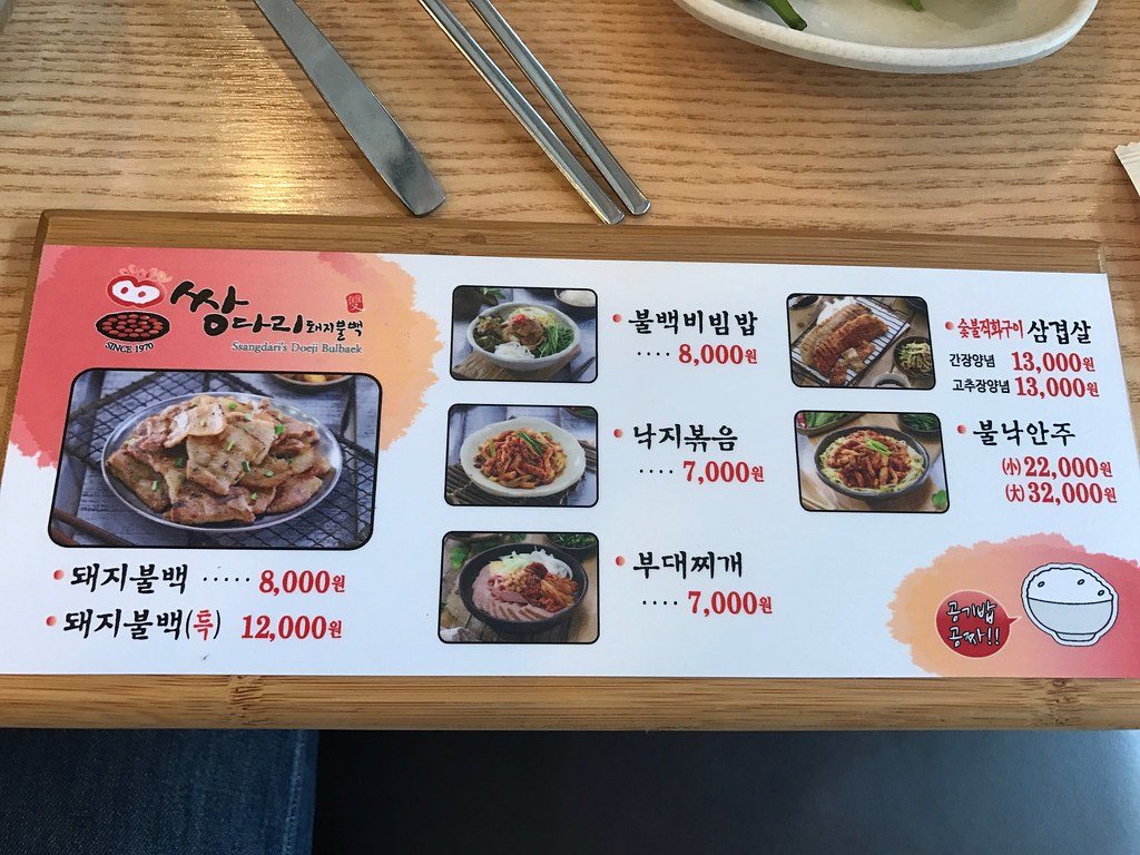 A menu board on a wooden table at Ssangdari Dwaeji Bulbaek listing 돼지불백 at 8,000 won and the special at 12,000 won, with photos of bulbaek bibimbap, stir-fried octopus and army stew at 7,000–8,000 won, and a speech bubble saying rice is free, beside steel chopsticks and a spoon, April 2017.