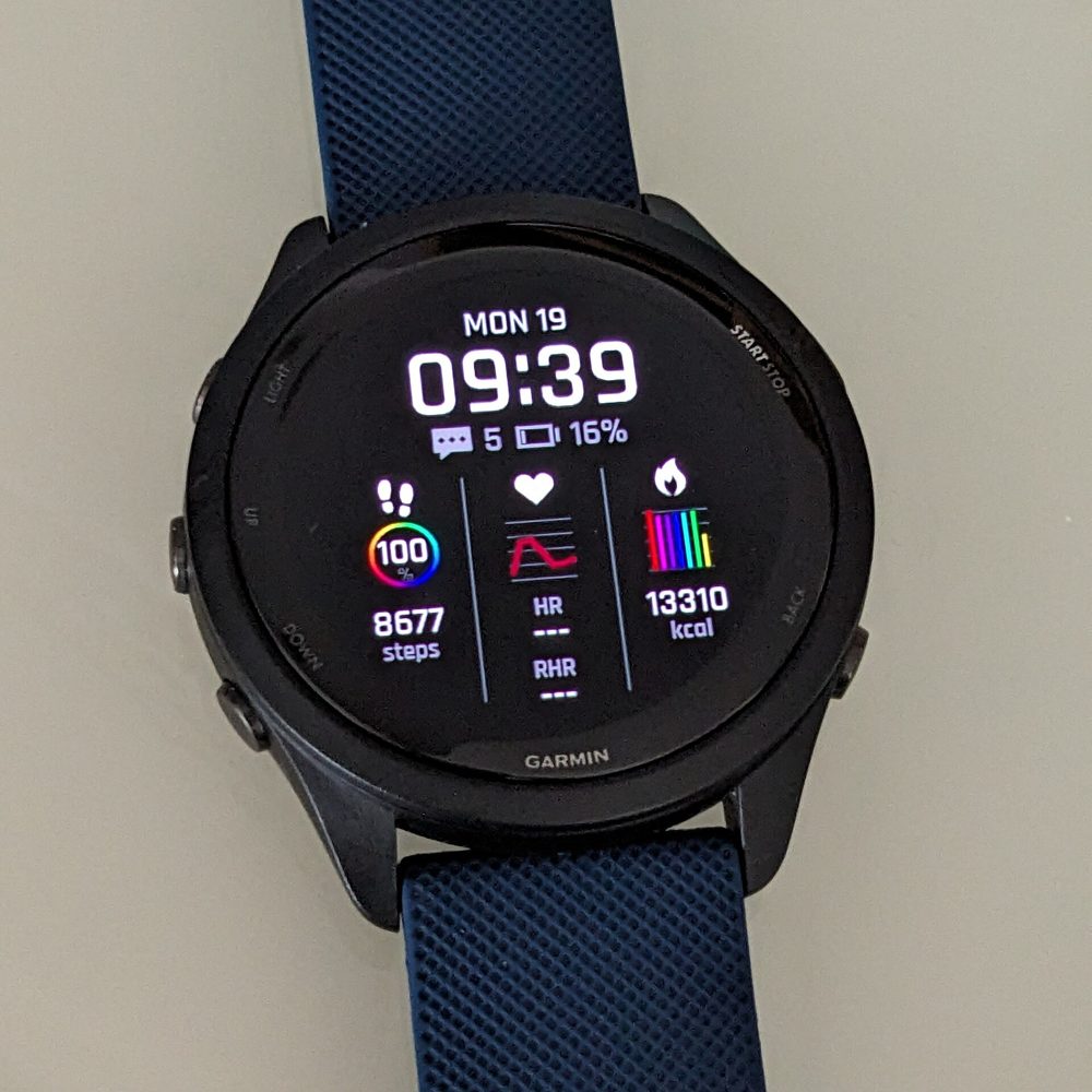 A Garmin Forerunner 265 running watch with a dark-blue strap lying flat, its round AMOLED face showing the time, steps, heart rate and calories.