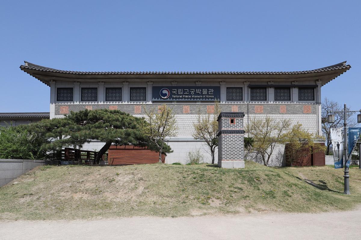 The National Palace Museum of Korea, a long white building with a traditional curved tiled roof, behind a low wall and pine trees under a clear sky.