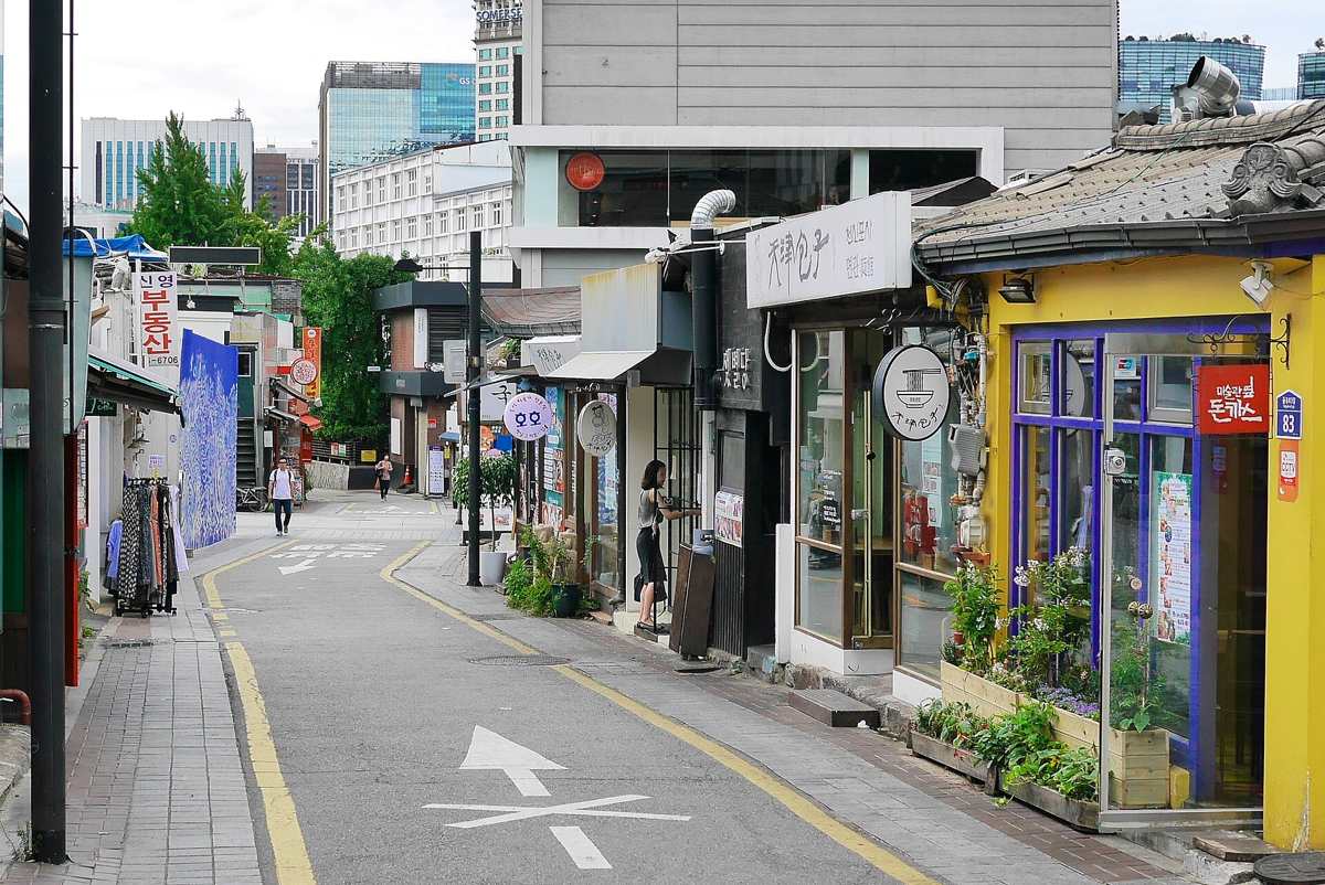 A narrow one-lane street in Samcheong-dong, Seoul, lined with small shops and a yellow-painted restaurant under a hanok roof, with narrow brick pavements on each side and a painted arrow on the asphalt.