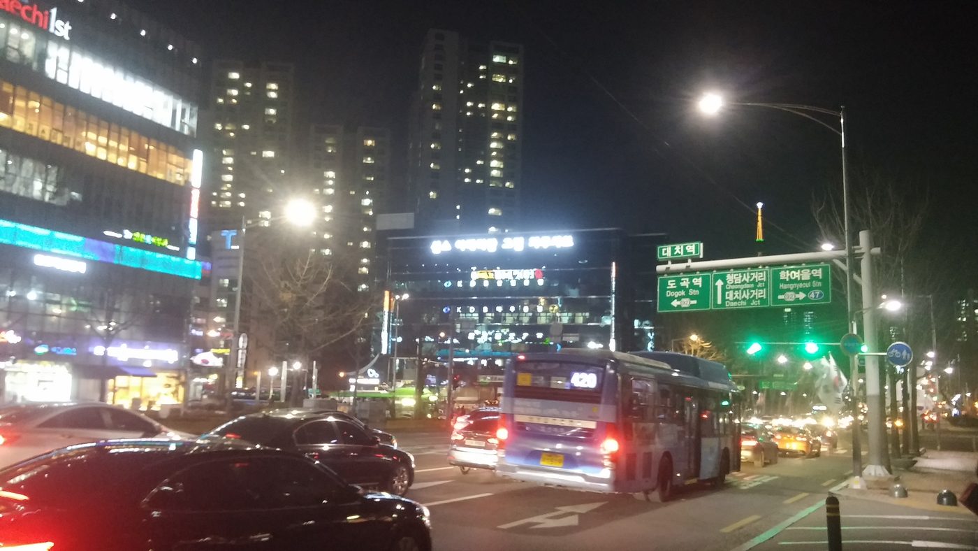 Daechi Station intersection in Gangnam at night: traffic lights, a green road sign marked 대치역 (Daechi Station), a bus and lit office and apartment towers, February 2016.