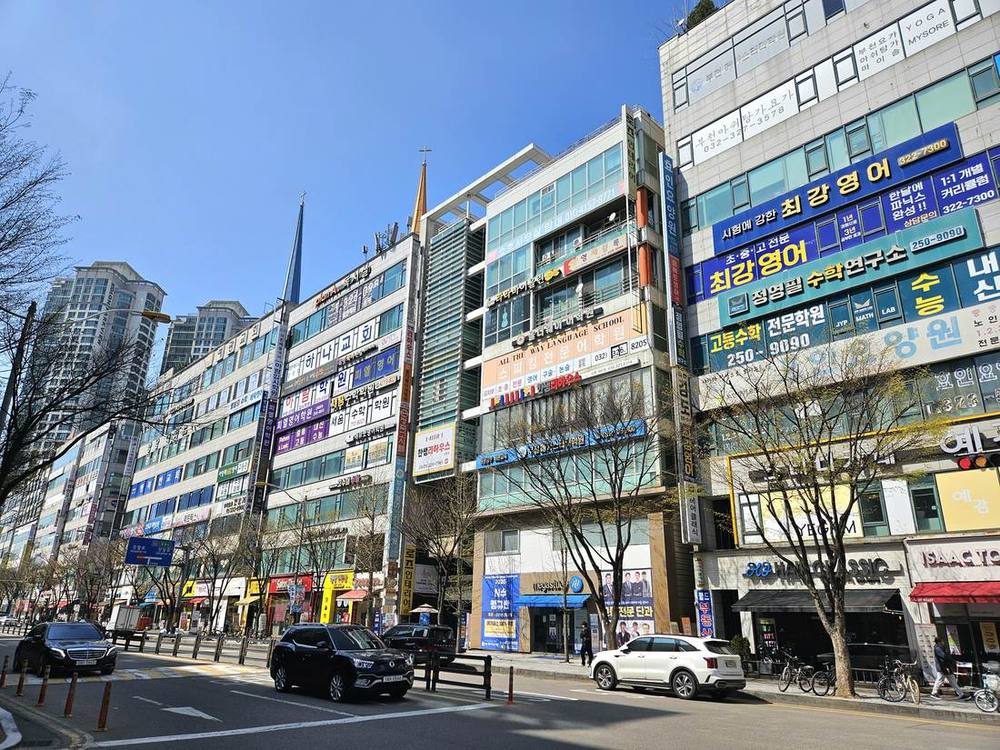 A street of mid-rise buildings in Sang-dong, Bucheon, covered in academy signs, including a large blue sign for 최강영어 (an English academy for elementary to high school), a green sign for a maths research institute, and a "high-school maths specialist academy" sign, under a clear sky in April 2025.