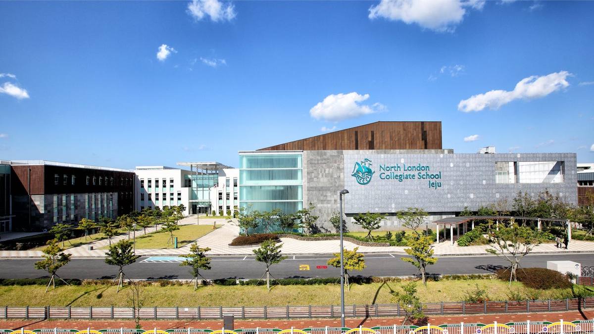 The front of North London Collegiate School Jeju: a long, low modern campus of glass, grey stone and brown cladding with the school's crest and name on the facade, behind a lawn and young trees, under a blue sky, March 2019.