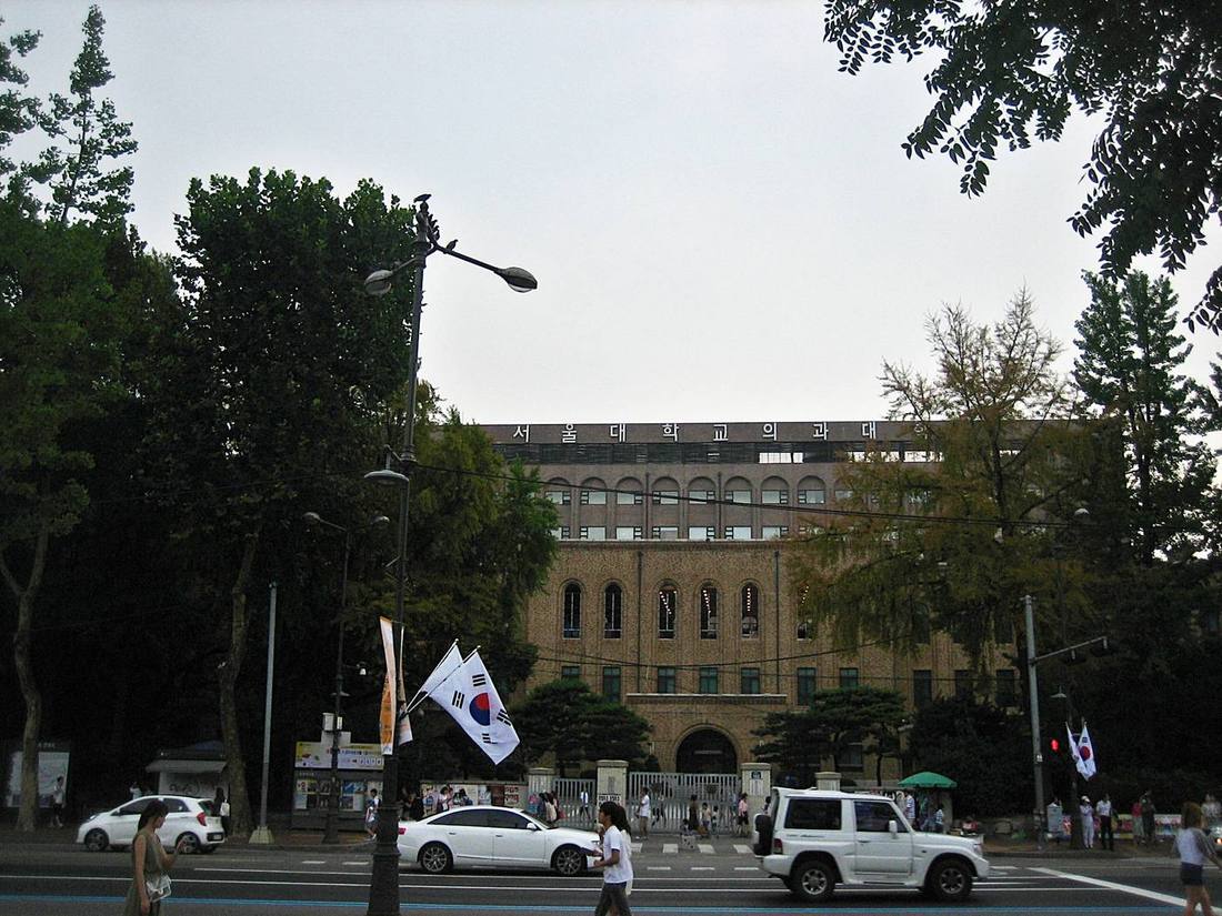 The brick façade of Seoul National University College of Medicine in Jongno, Seoul, with arched windows, the name 서울대학교 의과대학 across the top, Korean flags on lamp posts and cars passing on the road in front, August 2015.